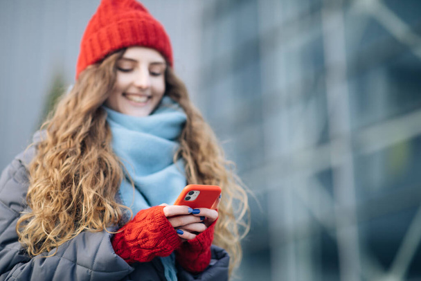Joyful female in good mood typing and scrolling on smartphone outdoors. Curly-haired female using smartphone standing outside. A smiling happy girl employee typing on a cellphone. Winter concept. - Fotó, kép