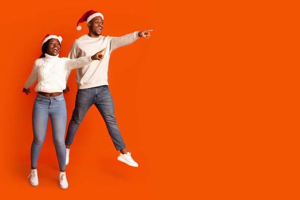 Look There. Joyful Black Couple In Santa Hats Jumping And Pointing Aside - Zdjęcie, obraz