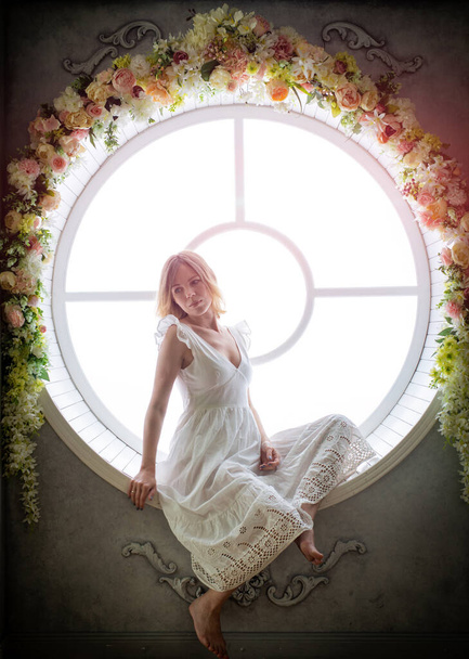 A woman in a white dress 35-45 years old sits in a round window. She looks away. White light comes from the window. - Fotoğraf, Görsel