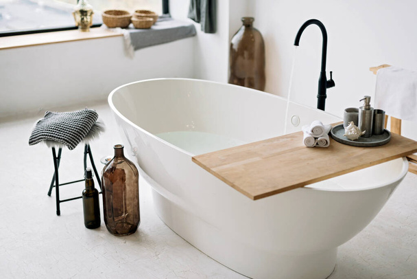 White bathtub fills with foam water in a modern apartment with stylish loft-style interior design, home decor. Spa concept, relaxation. Soft selective focus. - 写真・画像