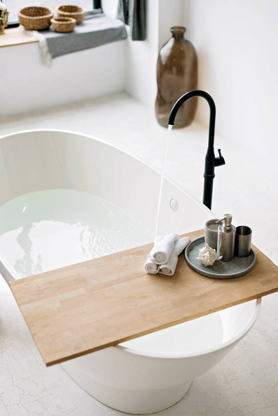 White bathtub fills with foam water in a modern apartment with stylish loft-style interior design, home decor. Spa concept, relaxation. Soft selective focus. - 写真・画像