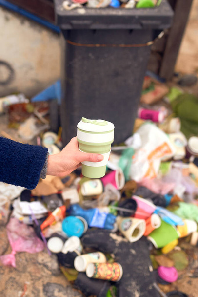Mano femenina sosteniendo eco taza reutilizable y un montón de basura en el fondo - Foto, Imagen