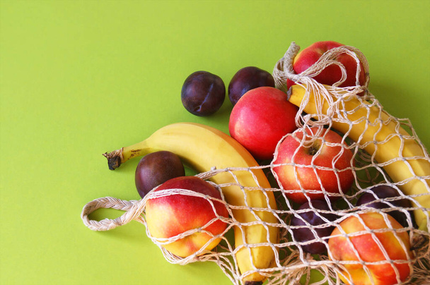 Красные яблоки, бананы, сливы и струны мешок на ярко-зеленом фоне. Свежие фрукты на столе и копировать пространство. Лето или осень. - Фото, изображение