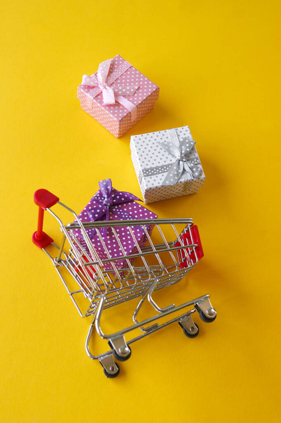 Sarı arka planda küçük hediyeler olan mini alışveriş arabası. Mağazalarda ve internette tatil için hediye alışverişi kavramı. - Fotoğraf, Görsel