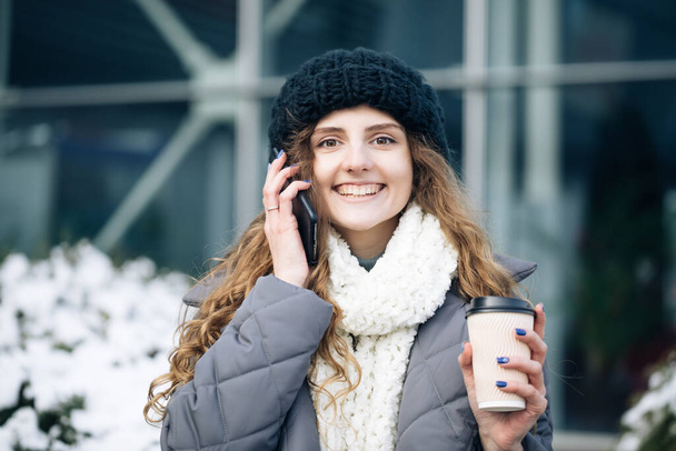 Curly haired woman using her smartphone and holding paper cup with coffee. Young girl talking on her mobile phone while walking in the city street - Foto, Imagem