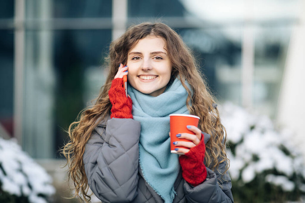 Curly girl talking on her mobile phone while walking in the city street. Curly haired woman using her smartphone and holding red paper cup with coffee. Woman walking and talking - 写真・画像
