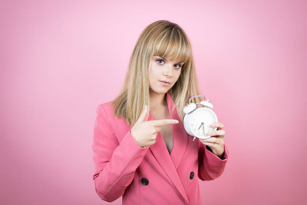 Молодая белая женщина на изолированном розовом фоне серьезно указывая на часы, потому что уже поздно - Фото, изображение