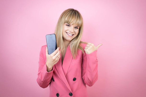 Молодая белая женщина на изолированном розовом фоне улыбается, показывая телефон и указывая на сторону - Фото, изображение