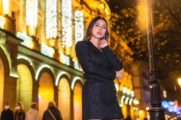 Christmas shooting, a pretty brunette girl in the city at night next to the Christmas lights of a beautiful building. Elegant black christmas or new year's eve dress with black boots - Foto, Bild
