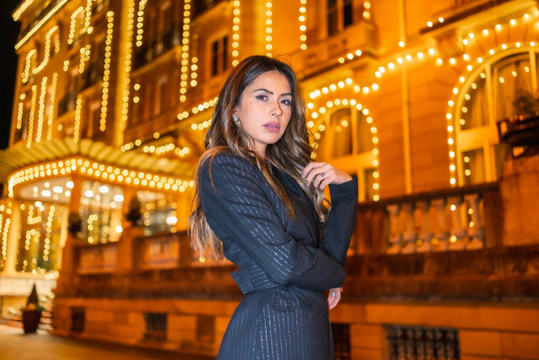 Christmas shooting, a Caucasian brunette girl in the city at night by the Christmas lights. Elegant black christmas or new year's eve dress with black boots - Foto, imagen