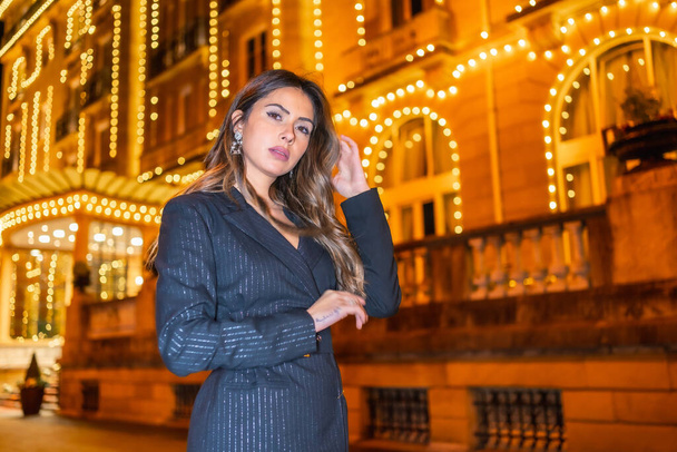 Christmas shooting, a Caucasian brunette girl in the city at night by the Christmas lights. Elegant black christmas or new year's eve dress with black boots - Fotografie, Obrázek