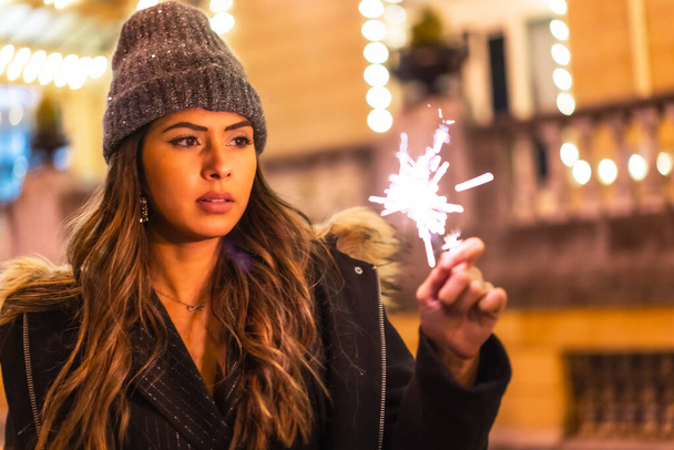 Christmas portrait with christmas lights in a beautiful building in the city, young caucasian woman at night with some torches in hand. With a winter hat - Fotografie, Obrázek