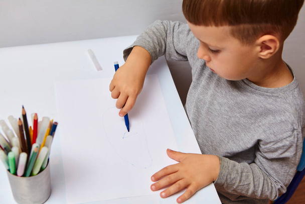 Sarı saçlı sevimli küçük çocuk evde renkli kalemler çiziyor. Beyaz masada çizer.  - Fotoğraf, Görsel