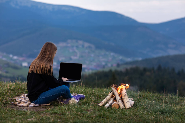 young female freelancer working on laptop in the mountains in the evening. Tourist girl sitting near campfire and having fun. Copy space - Photo, Image