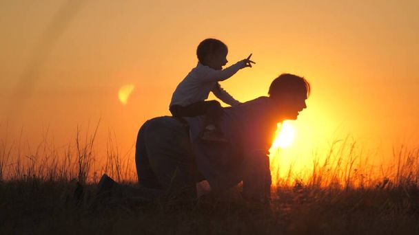 Папа играет со своей маленькой дочерью в парке на траве в лучах заката. Отец садится на спину, садится на папу, радуется. Папа и маленький ребенок играют на открытом воздухе. Счастливая семья - Фото, изображение