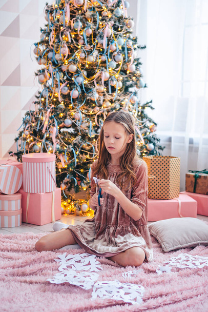 Ağacın yanında oturan ve kağıt kar taneleri yapan sevimli küçük kız. Oda dekore edildi. - Fotoğraf, Görsel