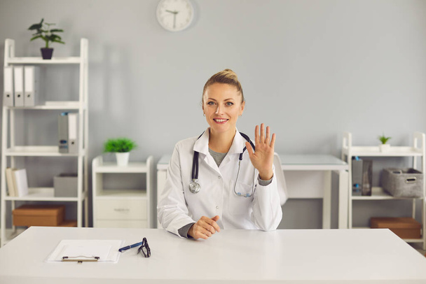 Young smiling blonde woman doctor nurse therapist sitting and greeting patient with hand online - Foto, imagen