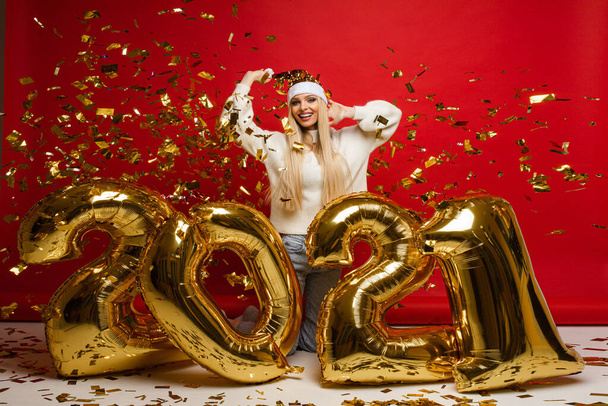 aantrekkelijke vrouw in comfortabele kleding in de buurt van grote gouden ballonnen met nummers 2021 en confetti - Foto, afbeelding
