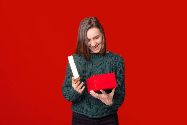 Удивлённая девушка открывает подарочную коробку и смотрит внутрь на красном фоне. - Фото, изображение