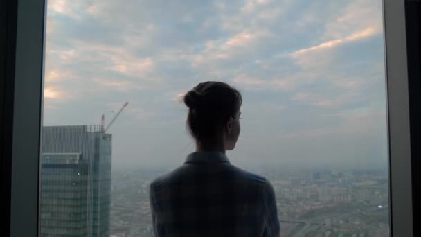 Πίσω όψη σκεπτικής γυναίκας που κοιτάζει το αστικό τοπίο μέσα από το παράθυρο του ουρανοξύστη - Πλάνα, βίντεο