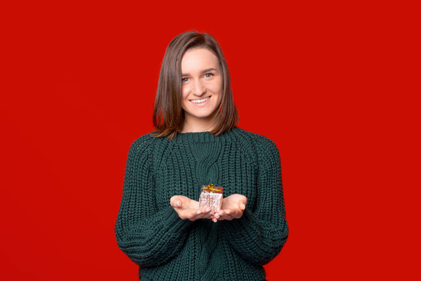 Усміхаючись на камеру, жінка тримає маленький загорнутий подарунок у руках на червоному тлі
. - Фото, зображення