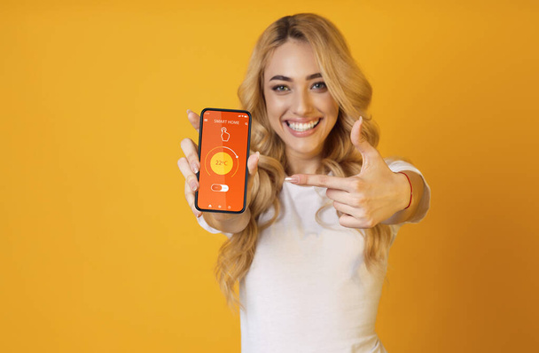 Jongedame wijst naar mobiele telefoon met smarthome app op het scherm, het regelen van de temperatuur over oranje achtergrond, collage - Foto, afbeelding
