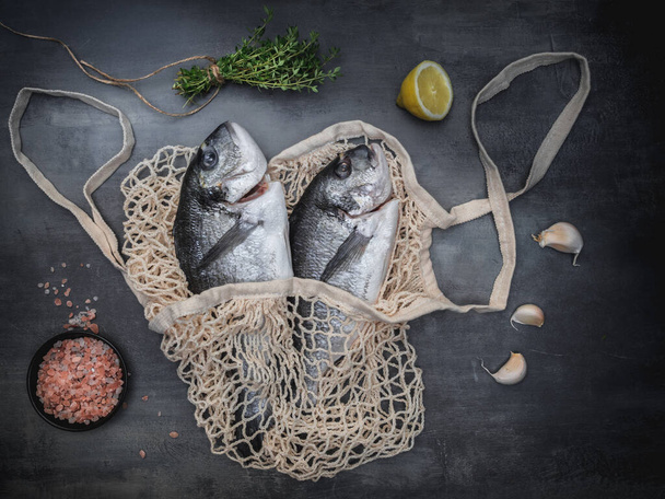Sea bream - dorado sea fish in shopping eco bag, thyme, lemon, garlic and Himalayan salt on dark vintage background. Overhead view with copy space. - Photo, Image