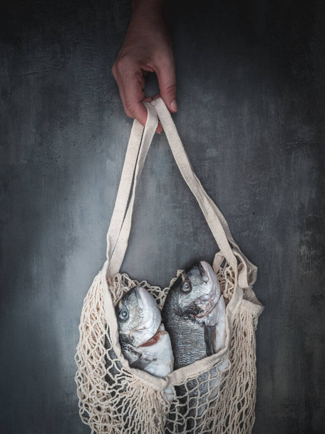 Mano femenina sosteniendo la bolsa ecológica de compras con dos besugo dorado pescado de mar. Cocina mediterránea. Vista aérea con espacio de copia. - Foto, Imagen