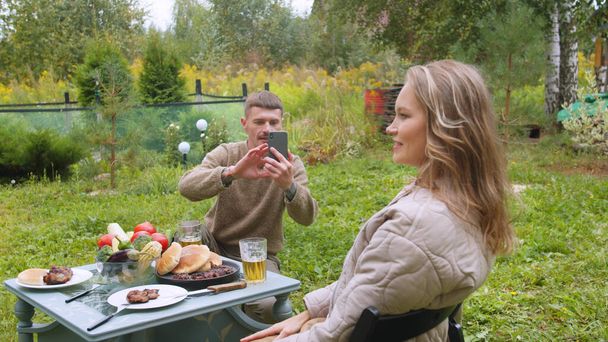 Муж фотографирует жену на смартфоне во время пикника в саду. Девушка позирует. - Фото, изображение