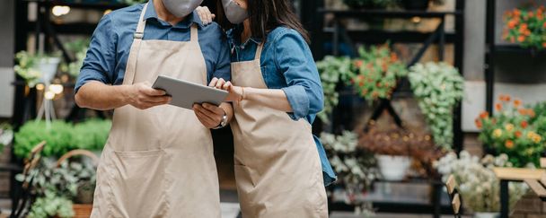 Young owners, man and lady in aprons and protective masks and look at tablet - Photo, Image