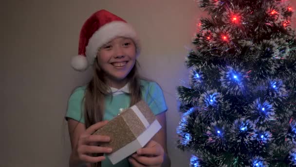 Ein kleines Mädchen mit Nikolausmütze fängt mit den Händen ein Geschenk neben dem Weihnachtsbaum. Kind mit Geschenkbox in der Hand, jubelt und lächelt. Familienurlaub für Kinder, Winterurlaub. Frohe Weihnachten. - Filmmaterial, Video