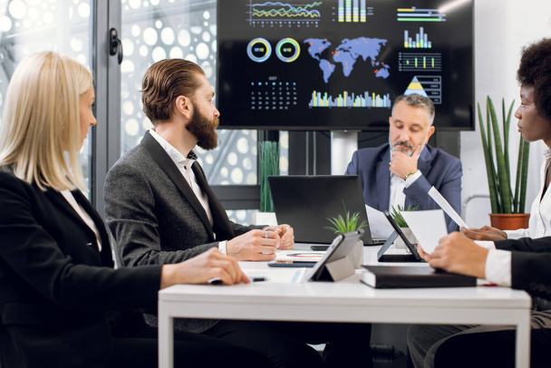 Team of multiracial concentrated businesspeople having meeting in boardroom at office, sitting at the table in front of a huge plasma TV screen. Focus on bearded man talking to colleagues - Photo, Image