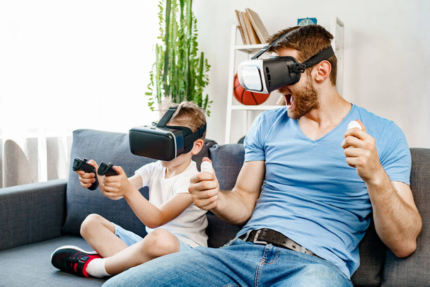Dad and son sitting on sofa and playing games with VR glasses - Foto, afbeelding
