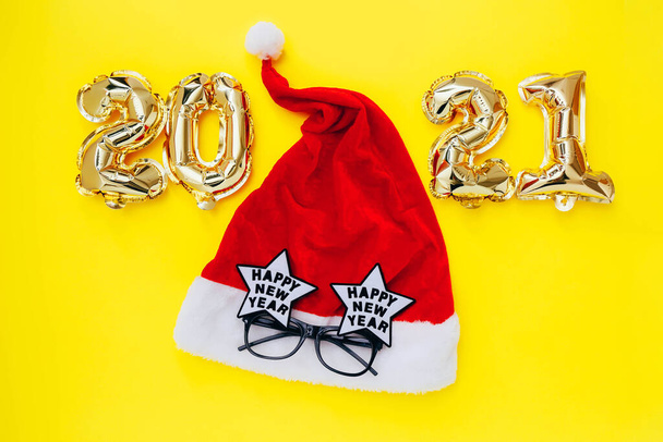 Fóliové balónky ve formě čísel 2021 a Santa klobouk se slavnostními brýlemi na žlutém pozadí. Novoroční oslava. - Fotografie, Obrázek