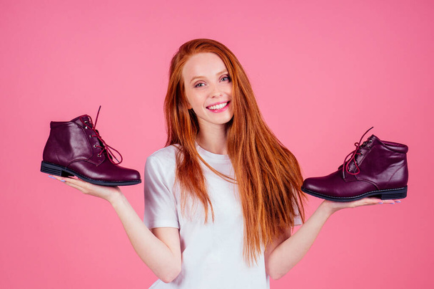 redhaired ginger woman in wtite cotton t-shirt holding burgundy lace-up boots in pink studio - Foto, Bild