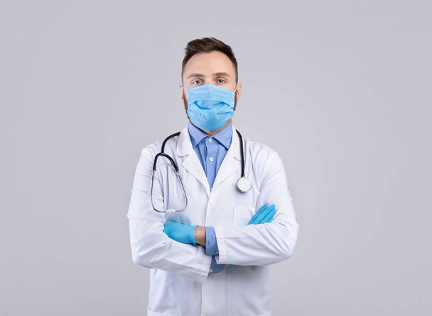 Szakmai férfi orvos egyenruhában, arc maszk és gumikesztyű pózol szürke stúdió háttér - Fotó, kép