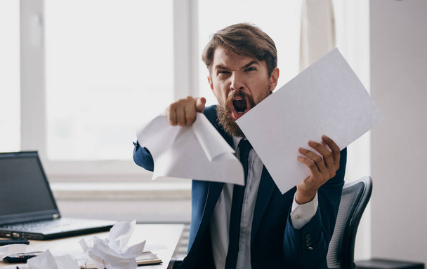 Homme d'affaires agressif dans un costume avec des feuilles de papier blanc dans ses mains dans le bureau près de la fenêtre - Photo, image