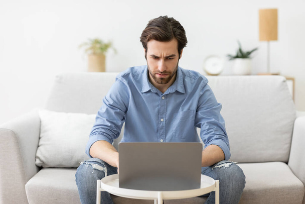 Mies Frowning käyttäen kannettavaa tietokonetta ottaa ongelma istuu sohvalla sisätiloissa - Valokuva, kuva