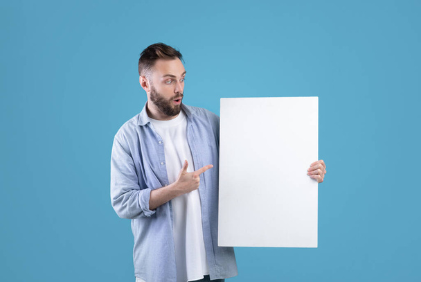 Έκπληκτος χιλιετής άνθρωπος δείχνει λευκό κενό αφίσα στο μπλε φόντο στούντιο, mockup για το σχεδιασμό αγγελία σας - Φωτογραφία, εικόνα