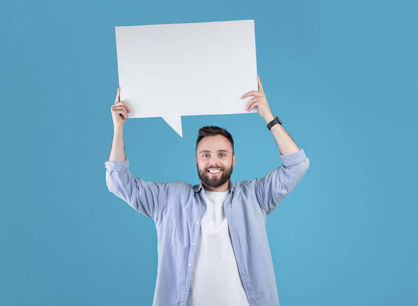 Happy millennial man holding blank speech bubble above his head over blue studio background, copy space - Photo, Image
