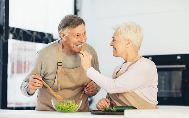 Coppia anziana che si nutre a vicenda e ride godendo cucinare all'interno - Foto, immagini
