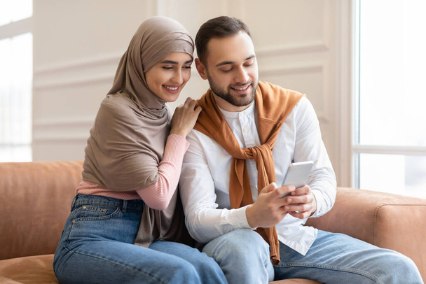 Молода мусульманська пара використовує мобільний телефон сидячи на дивані
 - Фото, зображення