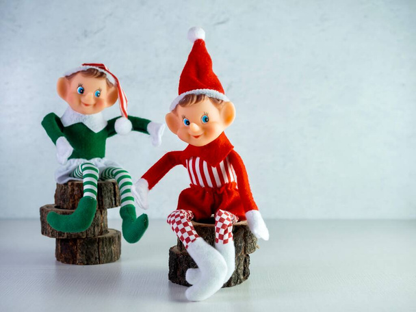 Dwa Elfy Boże Narodzenie siedzi na drewniane pnie kawałki, jeden w zielonym stroju i jeden w czerwonym stroju. Wystrój wakacyjny, Santa Hat, elf, czerwony i zielony. - Zdjęcie, obraz
