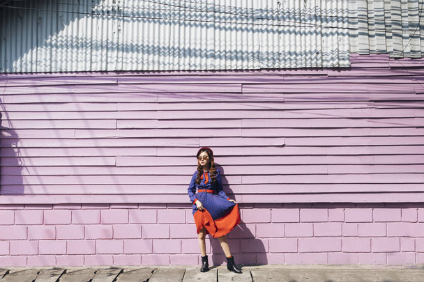 Wavy brunette woman in blue and red retro dress standing at the pink brick and wooden wall in the sunlight. - 写真・画像