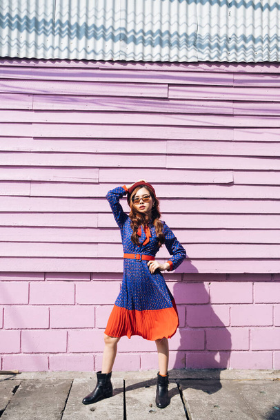 Vlnitá brunetka žena v modrých a červených retro šatech stojící u růžové cihly a dřevěné zdi se supermodelkou vnitřní na slunci. - Fotografie, Obrázek