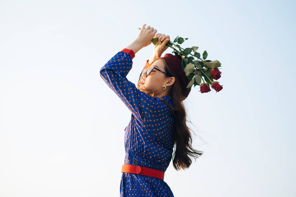 Winkelaufnahme einer welligen brünetten Frau in blau-rotem Retro-Kleid, die mit Rosen über dem Kopf im Sonnenlicht steht. - Foto, Bild