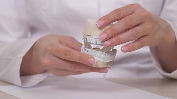Uma mulher ortodontista segura na mão uma placa para correção dos dentes e oclusão. Para colocar a placa sobre os dentes brancos de gesso. Tratamento da mandíbula em um consultório médico especializado. Um saudável e bonito - Foto, Imagem