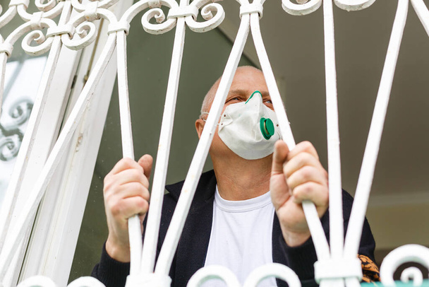 Карантин коронавируса. Старший мужчина в защитной маске за окном остаться дома, чтобы избежать заражения COVID-19 - ответственность и концепция профилактики - Фото, изображение