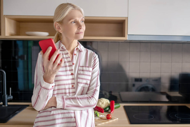 Nő áll a konyhában egy okostelefonnal a kezében, és néz figyelmes - Fotó, kép