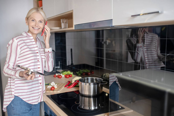 Woman standing near the stove in the kitchen and talking on the phone - Fotoğraf, Görsel
